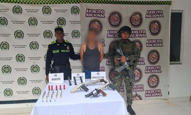 Hombre capturado, al lado un funcionario de la polic&iacute;a y al otro del ej&eacute;rcito con una mesa en frente con armas de fuego y municiones.