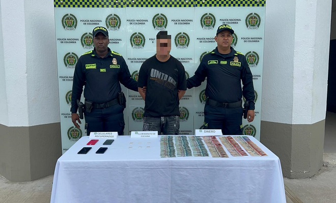 Hombre capturado por el delito de tráfico, fabricación o porte de estupefacientes con dos funcionarios de la Policía Nacional sujetándolo y brindando seguridad y sobre una mesa el material incautado. 