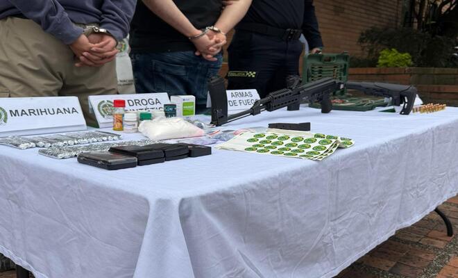 Mesa cubierta con mantel blanco donde se exhiben objetos etiquetados, incluyendo paquetes, frascos, municiones y armas largas; al fondo se observan personas con las manos sujetas detr&aacute;s del cuerpo.